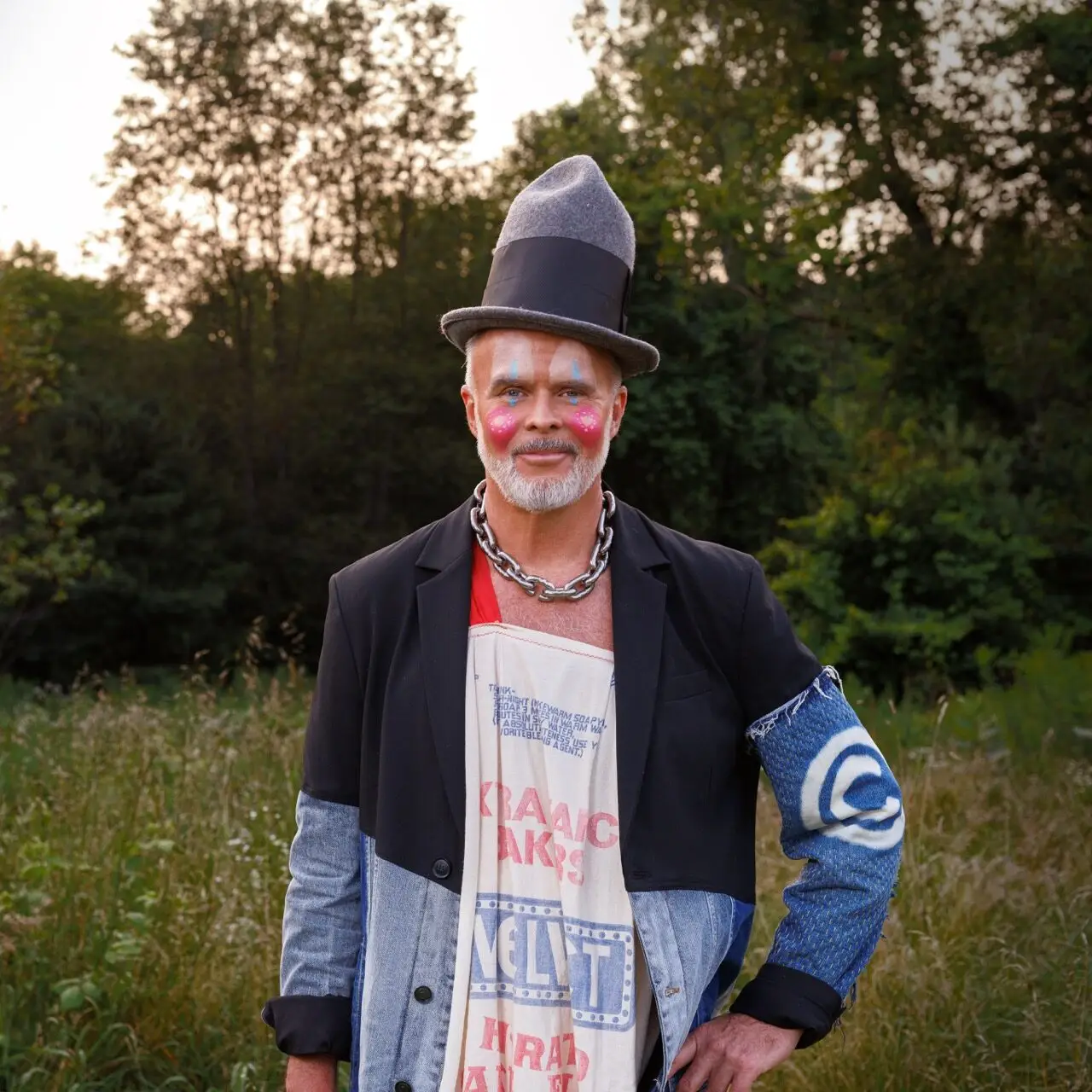 Barr Gilmore standing in a meadow in a tall felt hat, face paint and a hand-printed jacket