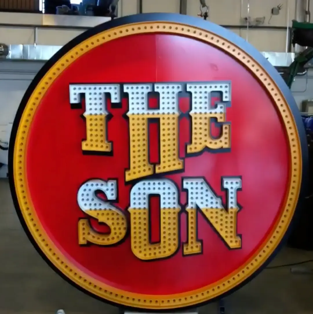 A circus-style illuminated sign spelling THE SON in gold bulbs on a red disc