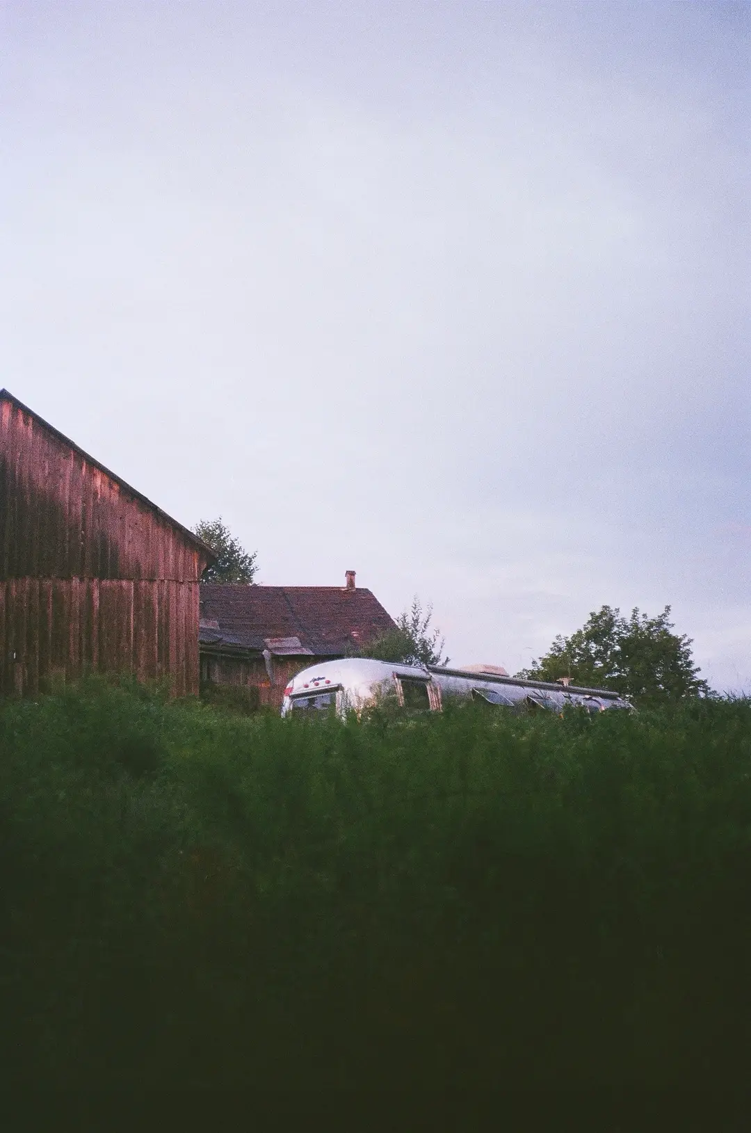 The weathered barn and the Airstream trailer standing in long grass under an overcast sky