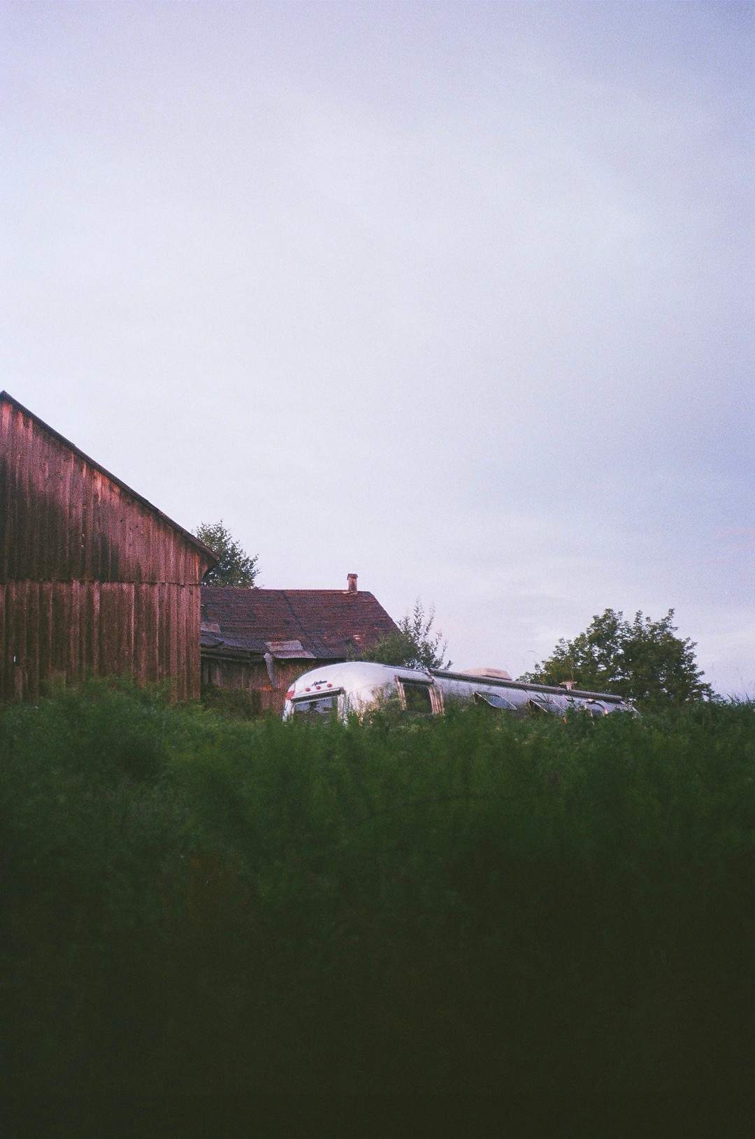 The weathered barn and the Airstream standing in long grass under an overcast sky